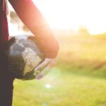 Global Phenomenon - person holding black and brown globe ball while standing on grass land golden hour photography